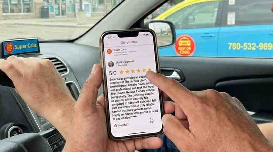 A person's hand with a slight scar holds a modern smartphone, displaying a Google Maps review of Super Cabs GP with a clear 5.0 star rating and review text. In the background through the car window, a matching yellow and blue Super Cabs GP minivan (cab 107, 780-532-9696) is visible. Diffused daylight fills the scene.